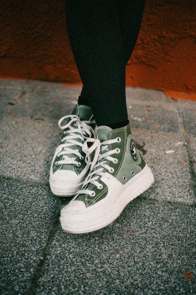 Green sneakers with white laces on pavement, captured outdoors with an orange wall backdrop.